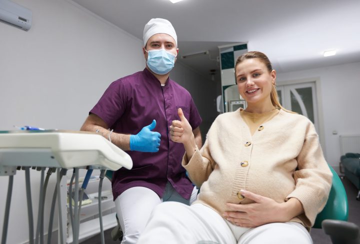 Dentist doctor and pregnant woman smiling a beautiful toothy smile, gesturing with thumb up at camera after regular dental check-up or teeth treatment in the dental office. People. Service. Healthcare