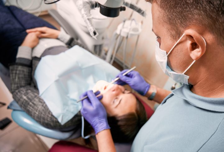 Close up of male dentist in medical mask checking patient teeth. Young woman lying in dental chair while having dental check-up in dental office. Concept of dentistry, stomatology and dental care.