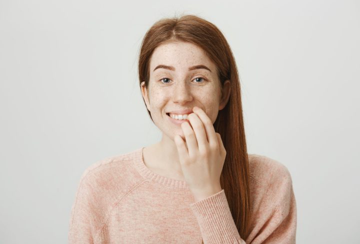 Close-up portrait of funny emotive caucasian ginger girl with bad habit, biting nails on fingers while looking at camera and standing against gray background. Woman is nervous during interview.