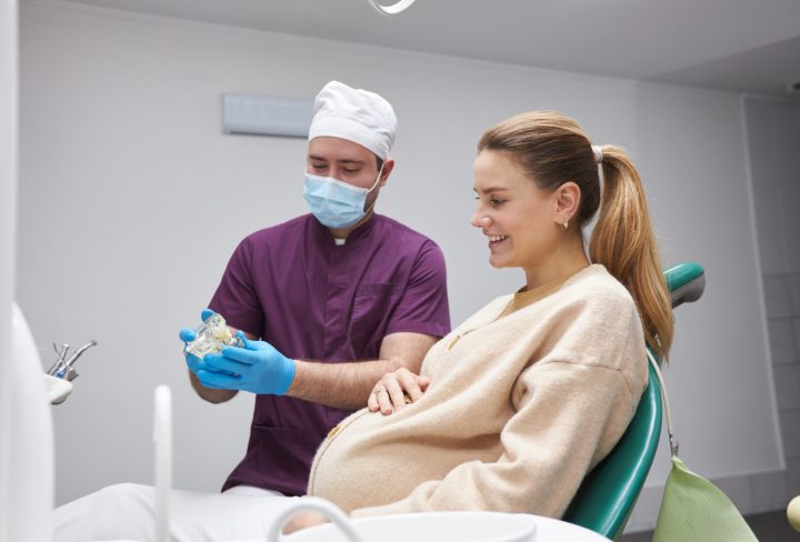 Pregnant woman cutely smiling while sitting in dentist's chair and listening to the doctor explaining to her on a model of human jaw bone, the treatment that needs to do in the dentistry clinic